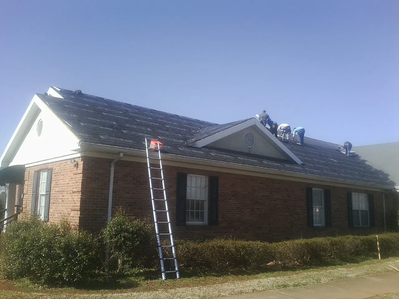 Crew tearing shingles off a brick ranch roof with a ladder set — Emergency Roof Repair work by our crew in Lansing